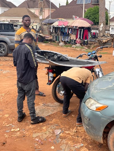 In Karshi, Nigeria, two men are working on a motorcycle and a damaged blue car. One man, wearing a black jacket with a distinctive pattern on the shoulders, stands facing the camera. A second man, wearing a tan shirt and black pants, is bent over, apparently working on the motorcycle or the car. The front of the motorcycle has been removed and is leaning against the bike. The car's front bumper is damaged. They are on a dirt road with scattered debris. In the background, there is a display of clothing under an umbrella, a parked gray pickup truck, and various buildings.