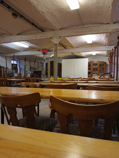 This image captures an empty, large multi-purpose room located in Homburg, Germany, configured with seating for a gathering or presentation. The room contains numerous long, light-colored wooden tables arranged in rows, each accompanied by wooden chairs with dark, patterned fabric seats. At the far end, a large white projection screen is centrally positioned, with a white projector resting on a table in front of it. To the right of the screen, an antique-style wooden display cabinet with glass doors contains various items.

The ceiling is low, rough, and light-colored, exhibiting exposed beams and visible grey piping. Multiple rectangular fluorescent light fixtures are recessed into the ceiling, providing illumination. A tall, silver-colored standing patio heater with a red glowing top is positioned in the mid-ground, emitting a reddish light. A prominent, light-colored cylindrical metal column supports the ceiling in the mid-ground. Along the left side of the room, a structure consisting of red-painted metal pipes with several suspended chain-and-hook mechanisms is visible. The floor is a light-colored concrete or stone surface. No individuals are present in the scene.