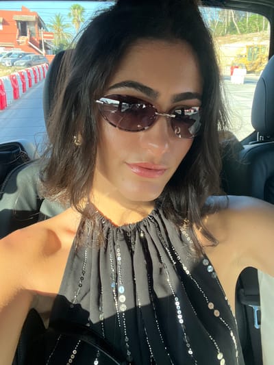 A woman with dark hair and sunglasses is in the passenger seat of a vehicle, with her head tilted towards the camera. She is wearing a black halter top with sequins. Outside the car, palm trees and buildings are visible, along with construction equipment. The scene appears to be in Santa Eulària des Riu, Spain.
