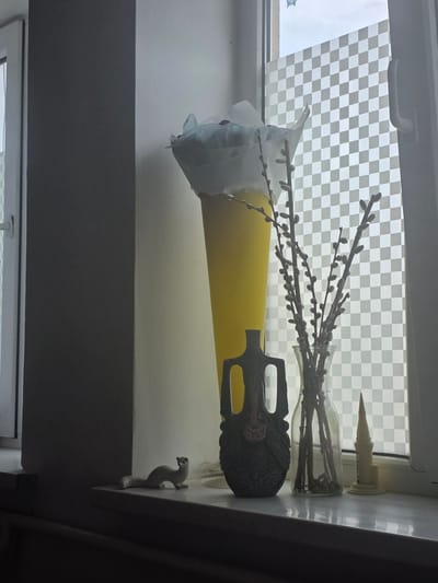 The image depicts a windowsill in what appears to be an interior setting, possibly a home in Novopokrovka, Kyrgyzstan. The windowsill itself is white and reflective. On the windowsill are several objects: to the left, a small white figurine of an animal, possibly a stoat or weasel, is positioned facing right. Next to it is a dark, ornate vase or vessel with a textured surface and handles. To the right of this vase is a clear glass vase containing several branches of pussy willow. To the right of the pussy willow, there is a small, light-colored, spire-shaped object resembling a miniature tower or candle holder. Behind these objects, a tall, yellow conical shape is visible, wrapped partially in white paper or fabric. The background shows a window with a checkered privacy film. The lighting suggests it is daytime, with light coming from the window, casting shadows on the wall to the left. There are no people visible in the image.