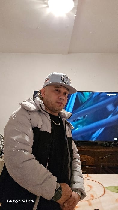 An adult male is depicted indoors, framed from the waist up. He possesses light brown skin, short dark hair styled in dreadlocks or braids visible at the sides, and a close-cropped beard. He wears a light grey baseball cap adorned with multiple dark emblems, a light grey and black puffer jacket over a black shirt, a silver-colored chain necklace, and a dark wristwatch on his left wrist. His hands are clasped in front of his torso, and he maintains a neutral expression while looking directly at the camera. The background consists of light-colored walls and a white ceiling featuring a bright, circular light fixture. To the viewer's left, a portion of a black and white circular fan is visible. Behind the subject, a large flat-screen television displays abstract blue and green visuals, with text that appears to be "waydread" or similar in the upper right corner of the screen. A wooden entertainment unit is positioned beneath the television. A light-colored patterned blanket or rug with a brown circular design is visible on a surface in the lower right foreground. The indoor environment provides no discernible visual indicators to confirm a specific location within Real Sitio de San Ildefonso, Spain.