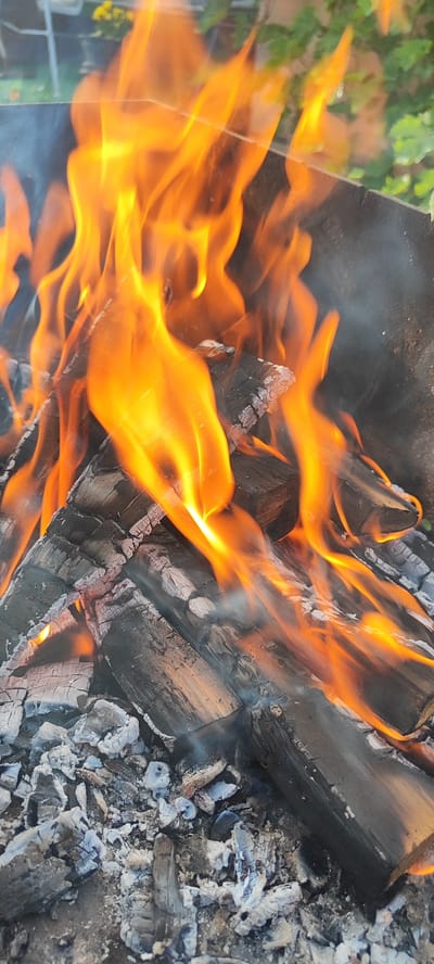 A close-up, outdoor, vertical shot depicts a bonfire with vibrant orange flames licking and engulfing charred logs and white ash in what appears to be a yard in the Lioznenski selski Savet region of Belarus. The wood is heavily burnt and appears to be in various stages of combustion, with some pieces still intact and others reduced to glowing embers. Smoke drifts upwards and to the left, obscuring parts of the background which shows a hint of green foliage and possibly a fence or outdoor furniture, suggesting a domestic or recreational setting. The focus is on the intensity and movement of the fire, highlighting the textures of the burning wood and ash. There are no people or animals visible in the image.
