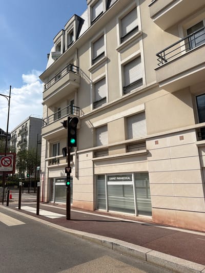 The image shows a street scene in Châtenay-Malabry, France, featuring a modern building with a facade of light-colored stone and beige stucco. The building has multiple stories, with balconies on the upper floors. There are several windows with closed roller shutters. On the ground floor, there are two storefronts, one with a sign that reads "CABINET PARAMEDICAL". A traffic light is visible, displaying a green signal. Adjacent to the traffic light is a sign with a circular red border and a diagonal red line across a symbol that likely indicates a prohibition, possibly "No Entry" or "No Stopping". Another smaller sign below it appears to be a bicycle crossing symbol with a green pedestrian figure above it. The street in the foreground is paved, with a crosswalk marked by white stripes. A few dark bollards are positioned along the sidewalk. The sky is blue with some clouds. The overall environment suggests a daytime scene in an urban or suburban setting.