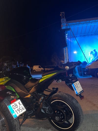 The image depicts an outdoor night scene in Gorna Rositsa, Bulgaria, featuring two motorcycles parked on an unpaved surface in the foreground, with an active concert stage in the background. The motorcycle on the left has a red rear fender and a Bulgarian license plate "OB K 1956 BG" with "BG" and the European flag emblem. The motorcycle to its right, primarily in the mid-ground, is a black and neon green/yellow Kawasaki Z-series, with a Givi branding visible near the rear seat. Its rear tire features a white reflective stripe, and its Bulgarian license plate reads "EB K 1710 BG", also bearing the "BG" and European flag emblem. The ground beneath the motorcycles is dirt or gravel.
In the mid-ground to the left, a small group of approximately 3-5 individuals is partially visible and blurred, observing the scene. Further back, two white vans and at least one other parked vehicle are discernible on the unpaved lot.
The background is dominated by a brightly illuminated outdoor stage, constructed with metal truss scaffolding, bathed predominantly in strong blue stage lights, creating a noticeable haze or smoke effect. Multiple figures, presumably band members, are visible on stage, with at least two distinct individuals on the right side. Musical equipment, including microphones and speakers, is present. A bright, central light source on the stage casts a strong glare. A prominent diagonal cable or guy-wire stretches across the upper portion of the frame, above the stage structure. The sky is dark, characteristic of nighttime.
