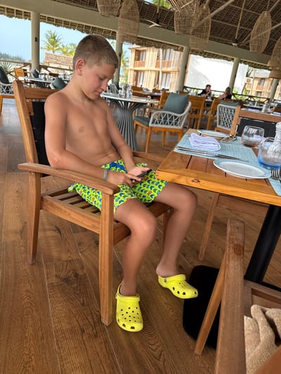A young male is seated in an open-air dining establishment in Michamvi Kae, Tanzania. The subject, appearing pre-teen, has short light brown hair and is shirtless. He wears neon green swim shorts with a repeating pattern of blue shapes and bright yellow Crocs. A silver-colored bracelet is on his left wrist. He is positioned in a light brown wooden chair with black strap-like backrests, looking down intently at a black smartphone held in both hands. To his immediate right is a light wooden square table set with a light blue placemat, a folded white napkin, silverware, an empty white plate with a blue rim, and sunglasses. A clear glass bottle and a textured light blue glass are also on the table. The floor is composed of polished wooden planks. In the background, numerous other dining tables and chairs are visible within the same open-air structure, which features a high, vaulted thatched roof with hanging woven pendant lamps. Beyond the restaurant's open side, multiple multi-story resort buildings constructed from natural materials, along with lush green tropical foliage and palm trees, are visible under a bright sky. Other patrons are seated at distant tables.
