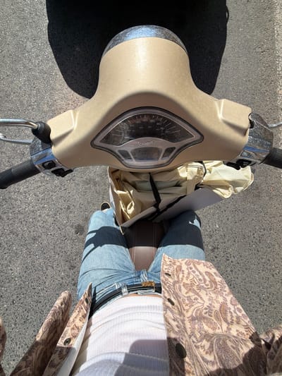 An overhead shot captures a person seated on a beige Vespa scooter, likely in Ondara, Spain, under bright, clear sunlight as indicated by strong shadows on the asphalt ground. The scooter's front fairing and handlebars are prominent on the left, featuring a speedometer with visible numbers (0-80) and the "Vespa" brand name partially seen below the dial.

The person is wearing light blue jeans, a white ribbed top, and a patterned jacket with brown and cream paisley or floral motifs. Their legs are positioned over the scooter's seat, which appears to be dark brown. A gold buckle is visible on their belt. A light-colored, possibly cream or off-white, folded item or bag is secured between the scooter's front and the rider, near the handlebars. The ground is dark gray asphalt, suggesting a street or parking area. The strong sunlight casts distinct shadows of the scooter and the person, reinforcing the sunny weather conditions.