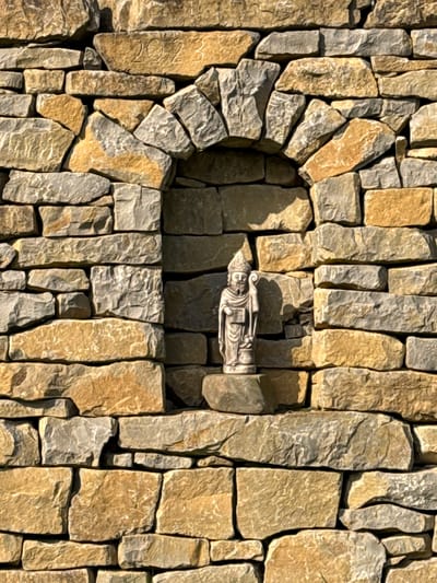 This image presents a close-up view of a dry-stacked stone wall located in Mertert, Luxembourg. The wall is constructed from irregularly shaped, rough-hewn stones, predominantly in shades of beige and grey. Centrally positioned within the wall structure is an arched niche. Inside this niche, a small, light-colored statue stands on a flat stone ledge. The statue depicts a bearded male figure adorned with a pointed hat or mitre, holding a pastoral staff in his left hand and a rectangular object, possibly a book, in his right hand. A stone block positioned above the apex of the arched niche, slightly to the right, is visibly inscribed with the numerals "2023". The scene is illuminated by direct sunlight, casting distinct shadows that highlight the texture and three-dimensional relief of both the stonework and the statue. No human figures or dynamic actions are present.