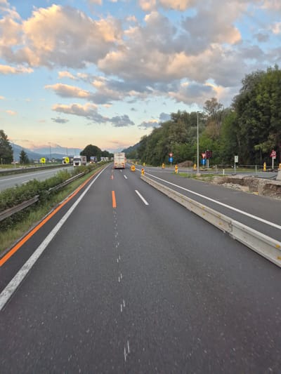 The image depicts a highway in Weer, Austria, during what appears to be late afternoon or early evening. The sky is filled with dramatic clouds, tinged with pink and orange from the setting sun, juxtaposed against a clear blue sky. The highway is asphalt, with clear lane markings including a solid white line and a dashed orange line marking the lane. There is a concrete barrier separating the lanes.

On the highway, several trucks are visible, traveling in the same direction as the viewer. Traffic appears moderate. The roadside is lined with vegetation and trees, with mountains visible in the distance. There are road construction signs and barriers present, indicating ongoing work or a lane closure. Specifically, there are orange traffic cones, temporary barriers, and signs indicating lane diversions and speed limits. One sign clearly displays "80", indicating an 80 km/h speed limit. Another sign shows a circular warning symbol with a diagonal line and an arrow pointing upwards, likely indicating a mandatory direction or lane usage.

The overall impression is one of a typical highway scene with some traffic and ongoing road maintenance, set against a scenic mountainous backdrop under a striking sky.