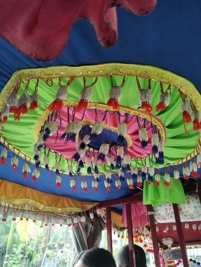The image depicts the interior of a decorated space in Southkhali, Bangladesh. The ceiling is draped with layers of colorful fabrics: blue, green, pink, and yellow. Tassels adorn the edges of the fabric layers, featuring white, red, and blue elements.

In the foreground, parts of two people's heads are visible. Outside, foliage indicates an outdoor setting. The overall impression suggests a celebratory or decorative environment.