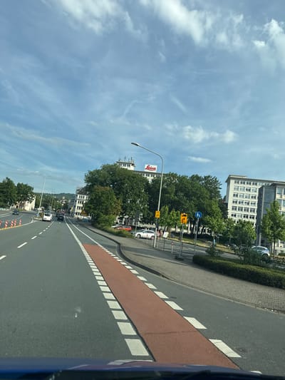 The image captures a view from a vehicle traveling along a street in Wetzlar, Germany. The scene is oriented vertically, with the sky occupying the upper portion of the frame and the road stretching into the distance.

In the foreground, a road with a distinct red bike lane marked with white squares is visible. The main lane of the road is grey asphalt with white dashed lines. Several traffic cones with orange and white markings are present on the right side of the road.

Along the side of the road, there is a pedestrian walkway paved with bricks. Beyond this, a row of green trees forms a barrier between the walkway and the buildings. Several buildings are visible, including a modern, multi-story white building with many windows on the left, and other structures further down the street. A tall lamppost stands prominently on the left side.

Traffic signs are visible, including a blue circular sign with a white bicycle symbol, indicating a cycle path. There is also a yellow sign with a black arrow pointing left, and another yellow sign with a black arrow pointing upwards. A red and white sign with "Leica" in red lettering is attached to a pole.

Several cars are driving on the road, including a white car in the distance and other vehicles further back. The sky is blue with scattered white clouds, suggesting a bright day. The lighting indicates it is daytime, likely late morning or early afternoon. The overall impression is one of an urban street scene during daylight hours.