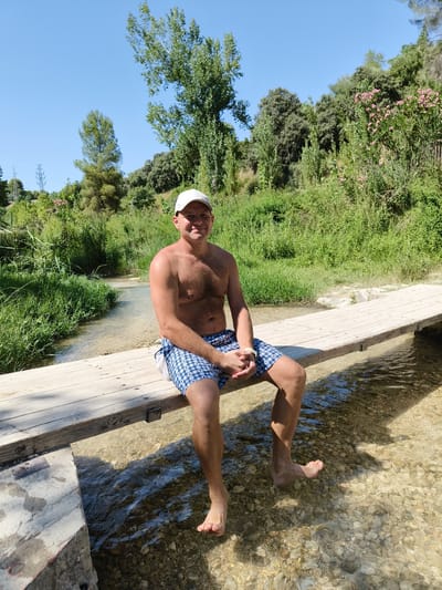 A shirtless man wearing blue and white checkered shorts and a white cap sits on a wooden footbridge over a shallow, clear stream in a natural environment, possibly a park or rural area. The man is looking towards the camera with a slight smile. His feet are dangling in the water, and his right foot is partially submerged. The stream bed is visible, with rocks and pebbles. Lush green vegetation, including trees and bushes, lines the banks of the stream. In the background, there are more trees, some with pink flowers, and a clear blue sky. The scene suggests a warm, sunny day.

The location is the Pou Clar in Ontinyent, Spain. This area is known for its natural springs and pools.