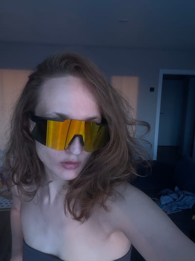 The image features a person's upper body and head, tilted to the side, in an indoor setting in Podshivalovo, Russia. The subject appears to be female, with light skin and long, wavy reddish-brown hair. She is wearing large, wrap-around sunglasses with highly reflective, golden-orange lenses and black frames, which obscure her eyes. Her lips are slightly parted. A dark-colored top or garment is visible on her shoulder and upper chest.
The background is dimly lit, suggesting evening or low light conditions. To the left and upper left, a plain, dark blue-grey wall is visible. Towards the upper right, there appears to be a lighter-colored architectural feature, possibly a doorframe or part of a built-in shelf, against a darker background. Further to the right, some crumpled, dark blue fabric or clothing can be seen on what might be a bed or couch. The overall atmosphere is muted due to the low lighting. There is no visible text in the image. The person is likely posing for a casual photo.