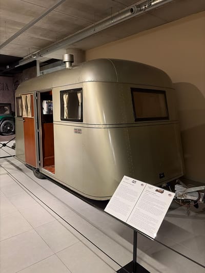 The image displays a vintage, streamlined travel trailer, identified by a nearby sign as a 1937 Pierce-Arrow Travelodge, within an indoor museum or exhibition setting. The trailer is a light grey or silver color, characterized by its riveted metal panels and rounded edges, which give it a distinct aeronautical appearance. It features multiple rectangular windows, one of which has curtains, and an open door that reveals a wooden interior. A metal exhaust or vent pipe protrudes from the side.
Next to the trailer, a white informational display board provides details about the "1937 PIERCE-ARROW TRAVELODGE" and also a "1927 FRANKLIN 11-B SEDAN." Visible text includes "LUCHTKOELING" (Dutch for 'air cooling'), "ENGINE 3-LITRE SIX-CYLINDER AIR-COOLED," and mentions of Franklin cars being "one of the first to" and having a "conventional design by J Frank DeCa Case."
The setting features concrete walls and ceilings with exposed metal piping, indicative of a modern exhibition space. The floor is covered with light-colored square tiles. Artificial lighting illuminates the scene. Partially visible beneath the trailer and through its open door are glimpses of other exhibits, including a wheel and the letters "CIA." There are no people visible. The Dutch text on the display board confirms a location within the Netherlands, aligning with the provided context of Wassenaar.