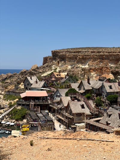 The image shows a coastal village with traditional wooden houses and a rocky cliff face in Mellieħa, Malta. The village appears to be a tourist attraction, with walkways, outdoor seating areas, and boats docked by the water. The architecture is rustic, with wooden facades and pitched roofs. The houses are clustered together on a hillside overlooking a bay. The sea is visible in the background, with a clear blue sky overhead. Some people are seen walking around the village and on the walkways. There are also several yellow kayaks and white jet skis on the dock. The overall scene is sunny and picturesque, suggesting a pleasant day for outdoor activities.