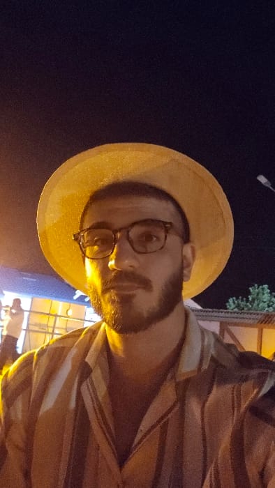 A single male individual is depicted in a close-up, chest-up portrait against a dark night sky in Dzoraghbyur, Armenia. The subject, appearing to be in his 20s to 40s, has a beard and is wearing prescription eyeglasses and a light-colored, wide-brimmed hat. He is dressed in a collared shirt with vertical stripes. A strong, warm artificial light source, likely from the left, illuminates his face and upper body. In the out-of-focus background to the subject's left, a blurred individual is visible near a blue-lit structure and light-colored framework. To the subject's right, dark green foliage from trees, a building with a reddish roof and patterned walls, and an overhead light fixture are discernible, indicating an outdoor urban or semi-urban environment, possibly an evening event or market.