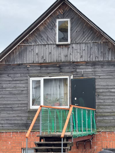 A photograph shows a gray, two-story wooden building with a gabled roof. The left side of the building has diagonally-laid wood siding, with a single small window. The right side has vertical wood siding. Above the right window is a black object. A makeshift balcony or platform covered with green netting extends from the right window, appearing structurally unstable. A brick building is visible to the right of the wooden structure. The image is taken from a low angle, with gray sky visible. There is no indication of the city of Ketoshi, Russia. There are no people in the image.