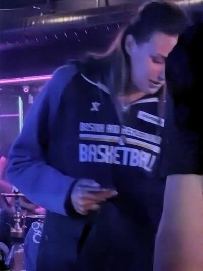 A person wearing a dark blue hooded sweatshirt with white and gold accents is standing indoors. The sweatshirt has text and a logo on the front. The person has dark hair pulled back and is looking down. The background is dark with some purple and pink lighting, suggesting a bar or club environment. There is no visible indication of the city of Travnik, Bosnia and Herzegovina in the image.