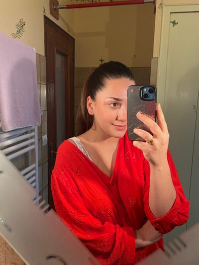 A female individual, aged approximately 20-30 years, is depicted taking a mirror selfie in a bathroom located in Ancenis, France. Her dark hair is pulled back into a ponytail. She is wearing a red, eyelet-embroidered, long-sleeved shirt, partially unbuttoned to reveal a white lace camisole or bra strap. She holds a dark-colored smartphone in her right hand, which obscures the right side of her face; her left eye and the left side of her face are visible, showing a slight smile. A gold ring with a small clear stone is present on her left ring finger. The background, reflected in the mirror, consists of light yellow walls adorned with green stenciled botanical and frog motifs. A dark brown wooden door is partially visible on the left. Above the door, a red cylindrical bar is mounted. A white, multi-barred towel warmer, over which a light purple towel is draped, is visible on the far left of the reflection. The mirror itself has an etched design along its bottom edge.