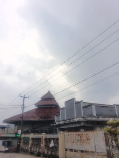 The image displays an urban street scene in Dakshin Hichachhara, India, under an overcast, light grey sky. A prominent building with a multi-tiered, pyramid-shaped roof clad in reddish-brown tiles and topped with a finial occupies the left-center. Its lower walls are light-colored. To the left, a dark utility pole supports several overhead power lines that traverse the upper frame. Adjacent to the pyramid-roofed structure, on the right, is a two-story grey concrete building with a flat roof and angular facade elements. The foreground features an ornate dark metal fence in front of the tiled-roof building, with a white textile fragment attached. To the right, a weathered, rusty metal gate serves as a boundary for the concrete building. A blurry white vehicle and a green traffic light indicator are partially visible on the far left. No individuals or specific actions are discernible.