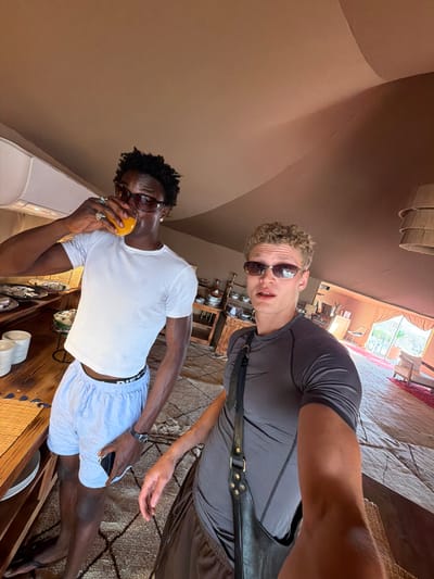 Two young men are posing for a selfie in a large, tent-like structure, likely a glamping tent, in Agafay, Morocco. The tent has a fabric ceiling and large windows or openings revealing an arid, desert landscape outside. The interior is furnished with wooden furniture, rugs, and seating.

The man on the left, with dark skin and curly black hair, wears a white t-shirt, light blue shorts, and sunglasses. He is holding a glass of orange juice to his lips. He has several rings on his fingers and a watch on his left wrist.

The man on the right, with lighter skin and curly blonde hair, wears a dark grey t-shirt, dark shorts, and sunglasses. He has a black bag slung over his shoulder. His expression is a slight grimace.

In the background, there is a wooden table with various plates and bowls, possibly from a buffet. Further back, a seating area with chairs and tables is visible, along with a view of the desert outside. The overall atmosphere suggests a luxurious desert retreat.