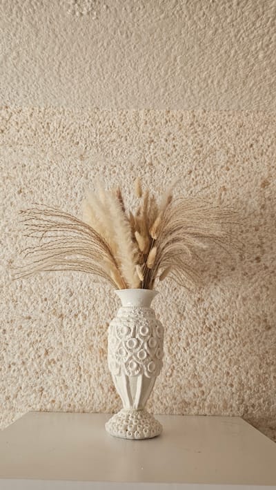 An ornate white vase containing dried pampas grass and wheat stalks sits on a white surface in front of a textured, cream-colored wall. The vase has a bulbous base and body, with a decorative pattern of circular motifs. The dried grasses are arranged in a fanned-out bouquet, with some stalks taller and more feathery than others. The lighting is soft and even, casting no harsh shadows. The background wall has a rough, pebbly texture, with a subtle horizontal line dividing it into two slightly different shades of cream. The overall aesthetic is minimalist and natural. The image was captured indoors, likely in a home or studio setting. There is no discernible location in Lubnya, Russia, or any other specific geographical context provided by the image.