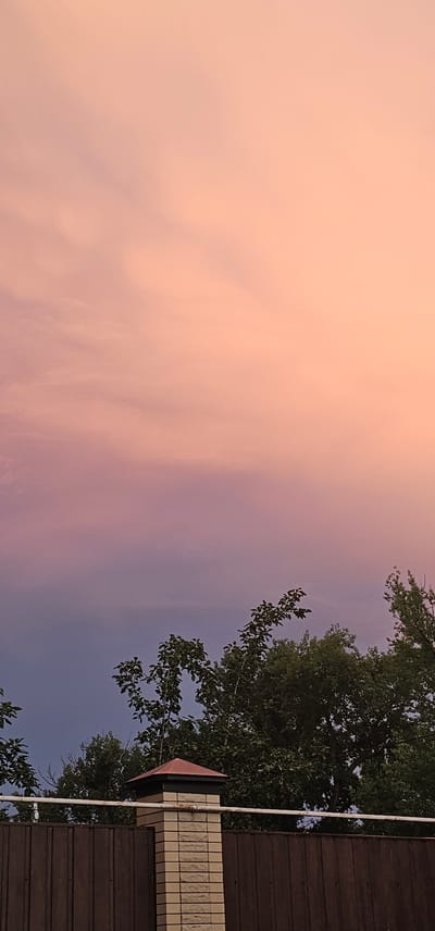 The image captures a serene outdoor scene likely at dusk, given the soft, gradient sky transitioning from a peachy pink to a dusky purple. The sky is largely the focus, with wispy clouds lending texture to the pastel hues. On the right side of the frame, a dark brown, corrugated structure, possibly a roof or siding, forms a vertical boundary. Adjacent to it, a chimney-like structure is visible, topped with light-colored, brick-like tiles and a pointed, reddish-brown cap. A white pipe runs vertically alongside the chimney. Lush green foliage from a tree overhangs the scene, its leaves silhouetted against the colorful sky. The overall mood is tranquil and atmospheric, evoking the quiet beauty of twilight in a residential or suburban setting. No people or text are visible in the image. The location context provided is "Овощной, Russia".