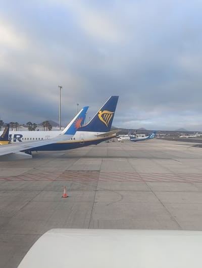 The image captures an active airport tarmac under an overcast, but bright, sky. The photograph appears to be taken from inside an aircraft, with part of a white wing or engine cowling visible in the lower right foreground.

The primary subjects are several airplanes parked on the concrete tarmac. Most prominently, the tail of a Ryanair aircraft (identifiable by its distinctive blue and yellow harp logo) is centered in the midground. To its left, another aircraft, likely also Ryanair, shows part of its fuselage with the letters "R" and "IR" visible, along with a blue tail featuring a bright orange sun logo. Beyond these, a blue and white Canaryfly aircraft is visible. Further in the distance, on what appears to be a runway, a white Malta Air plane is also parked.

The activity depicted is the routine ground operations of an airport, with aircraft parked, suggesting they are awaiting departure, arrival, or maintenance. No people are clearly visible, but ground staff might be present near the aircraft.

Notable details include the weather, which is predominantly cloudy with patches of lighter sky, indicating daylight hours, possibly morning or late afternoon. The landscape in the distance features arid, volcanic-looking hills or mountains, characteristic of the Canary Islands. Several palm trees are scattered around what appear to be airport buildings behind the closer aircraft. Tall light poles are visible on the tarmac. A single orange and white traffic cone stands on the concrete markings in the midground.

Visible text includes "RYANAIR" on the side of the closest aircraft, the "R" and "IR" on the adjacent plane, the full "CANARYFLY" logo on the blue aircraft, and "MALTA AIR" on the distant white plane. The unique liveries provide clear identification of the airlines. The scene reflects a typical airport environment in a warm, possibly island, climate, consistent with the San Bartolomé, Spain location (Lanzarote Airport).