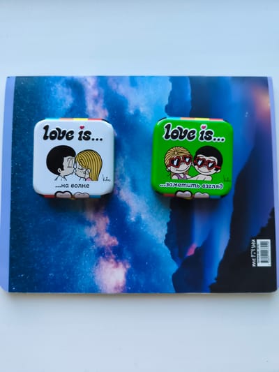 Two square, decorative tins are placed on top of a notebook with a starry sky or nebula design. The tin on the left is white with a drawing of a boy and girl kissing, with the text "love is..." and "...на волне" (which translates to "...on the wave") in Russian. The tin on the right is green with a similar drawing of a boy and girl wearing heart-shaped sunglasses, with the text "love is..." and "...заметить взгляд" (which translates to "...notice the glance") in Russian. Both tins have a rainbow-colored strip along the top edge. The notebook has a barcode and text on its bottom right corner. The image is taken from a top-down perspective. There is no identifiable information about the city of Bagerovo, Ukraine, in the provided image.