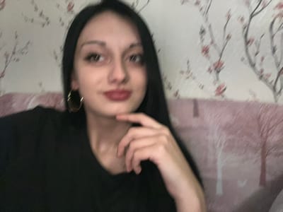 The image depicts an interior scene, stated to be in Lanchkhuti, Georgia, though visual evidence within the image does not confirm this specific geographical location. The primary subject is a female individual, appearing to be an adult, positioned centrally from the chest up. She possesses long, straight, dark hair parted on the side, dark eyebrows, dark eyes, a small nose, and lips colored with dark red lipstick, forming a slight smile or smirk. She wears a black v-neck top and a single gold-colored hoop earring is visible on her left ear. Her right hand is raised, with the index finger resting under her chin, adopting a thoughtful or posed gesture. The individual's gaze is directed slightly upwards and to her right (viewer's left). The background consists of a wall covered in light-colored wallpaper adorned with a pattern of dark, stylized tree branches and scattered pink or red blossoms. Below the wallpaper, a portion of a light pink or mauve upholstered furniture piece, possibly a sofa or headboard, with a textured or quilted appearance, is visible. The image quality is slightly soft or out of focus.