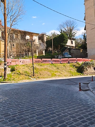 This image depicts an urban street scene in Drama, Greece, under a clear blue sky with scattered white clouds. The foreground features a dark grey cobblestone road surface, transitioning into an asphalt section further back. On the right, two orange and white striped traffic cones mark the edge of the road near a concrete curb and sidewalk element. A black utility pole with two spherical white lamps stands centrally, slightly to the left, with its shadow cast on the cobblestones. To its left, a tall, light brown wooden utility pole is visible, wrapped with white tape near its base.
Behind the street, a grassy embankment slopes upwards. A continuous line of approximately ten orange plastic construction barriers runs horizontally along the top of this embankment, separating it from the background. Behind these barriers, to the left, parts of a dilapidated, light-colored building with multiple windows and bare tree branches are visible. A section of a concrete wall with blue and white graffiti is present further to the right behind the barriers.
The background is dominated by a mix of buildings and vegetation. On the far left, a multi-story building with a light-colored facade and visible roof tiles is partially obscured by a bare deciduous tree. In the center-right, a crumbling, multi-story building heavily overgrown with dark green ivy is visible, featuring an open window on its upper floor. To the right of this, a stark, multi-story residential building with balconies and a plain, light-colored facade stands. Several bare trees with intricate branch patterns are scattered throughout the scene, one particularly large tree silhouetted against the sky in the upper mid-right. Overhead utility lines traverse the upper portion of the frame. No individuals are present in the scene.
