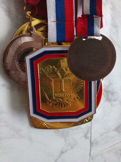 This is a close-up, eye-level photograph of several medals hanging from ribbons. The medals are a mix of gold and bronze colored, with red, white, and blue ribbons. One prominent gold medal features a double-headed eagle crest and the inscription "1 место," which translates to "1st place." Another medal, partially visible, appears to be bronze with a textured, ribbed design and a laurel wreath. A dark, circular object, possibly a third medal or a decorative element, obscures part of the gold medal. The medals are hanging against a lightly textured, off-white background, possibly a marble surface. The photograph does not provide any information about the specific location in the city of Polovinnoye, Russia.