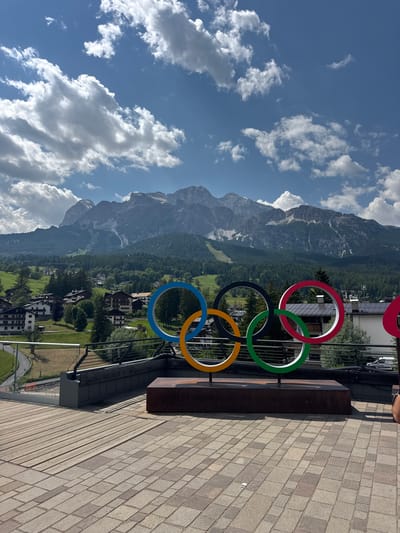 The image shows a prominent display of the Olympic rings, positioned in front of a breathtaking mountain landscape. The Olympic rings are rendered in their traditional colors: blue, yellow, black, green, and red. They are mounted on a dark, metallic structure.
The background reveals a vast mountain range, its peaks partially obscured by scattered white clouds against a vibrant blue sky. The lower slopes of the mountains are covered in lush green forests, transitioning to a charming village nestled in the valley below. The village comprises numerous buildings with traditional architecture, suggesting a European alpine setting. Green fields and trees dot the landscape, adding to the scenic beauty.
The foreground features a paved walkway made of rectangular stones, leading towards the Olympic rings display. A dark bench or seating area is adjacent to the rings. A sliver of a person's arm and hand is visible at the top right, suggesting a human presence, though no faces are clearly discernible.
The scene appears to be set during the daytime, with bright sunlight casting shadows. The weather seems pleasant, with a mix of sun and clouds. The overall impression is one of a majestic natural setting complemented by a significant cultural landmark, likely in Cortina d'Ampezzo, Italy, given the context. The image evokes a sense of grandeur and perhaps anticipation associated with the Olympic Games. No discernible text is present in the image.