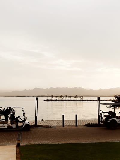 A photograph captures a serene outdoor scene in Ras Abu Soma, Egypt, at dusk or dawn, characterized by muted, sepia-toned lighting. The view features a body of water extending towards distant, hazy mountains, with a large, decorative sign reading "Simply Somabay" erected on a small landmass in the water. Two golf carts are situated in the foreground on either side of a paved brick area bordered by a patch of green grass. One cart is occupied by a figure in dark clothing, while the other is partially obscured, revealing dark seating and structural elements. Wooden planks form a pier or walkway extending into the water, with black poles marking its edge. The sky is overcast, contributing to the soft, diffused light across the scene.