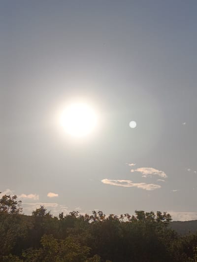 The image depicts a daytime scene, dominated by a bright, hazy sky. A bright sun is visible, and there is a fainter, smaller, circular shape alongside it that may be a lens flare. Scattered clouds appear in the lower portion of the sky. The bottom of the image features a tree line, with foliage in shades of green. The location, Barvinok, Ukraine, cannot be confirmed from the visual content.