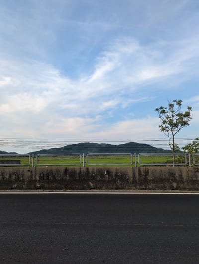 The image shows a roadside view with a concrete barrier, a metal fence, a green field, and mountains in the background. The sky is blue with scattered white clouds. The foreground features a dark asphalt road. In the mid-ground, a concrete barrier separates the road from a green field. A metal fence with a diamond pattern encloses the field. Utility lines are visible strung across the scene above the field. A solitary tree stands to the right of the field, its branches bare of leaves. The mountains in the distance are covered in green vegetation. The scene appears to be in an rural or semi-rural area. Based on the prompt requesting information about Kỳ Anh Commune, Vietnam, this image could depict a location along a highway or main road within or near this commune, characterized by agricultural land and distant hills or mountains. There are no people or animals visible in the image.