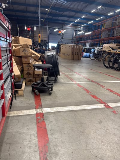 This is an interior shot of a warehouse in Sint-Baafs-Vijve, Belgium. The flooring is concrete with red and white painted lines indicating pathways. In the foreground and mid-ground, there are stacks of cardboard boxes, some with hazard symbols. A black rolling stool is positioned in front of a large, black cylindrical object. To the right, several bicycles are parked in a row. In the background, tall shelving units filled with more boxes are visible. A person is standing near the bicycles, facing away from the camera. The ceiling has exposed beams and industrial lighting.