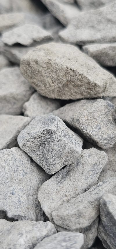The image shows a close-up, macro view of a pile of gray, angular stones. The stones vary in size and shape, with some appearing rough and others smoother. They are covered in a fine, sandy dust, which gives them a matte appearance. The lighting is even, suggesting it might be daytime with overcast skies, or taken indoors with artificial lighting. There are no people, animals, or any other living beings visible. The setting is simply a collection of rocks, likely aggregate or gravel, with no discernible background or location cues beyond the texture and color of the stones. No text is visible in the image.