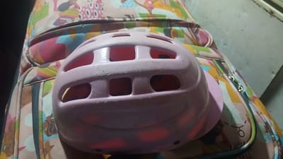 The image shows a pink helmet resting on top of a colorful patterned suitcase. The helmet has several rectangular openings for ventilation. The suitcase has a busy pattern with various shapes and figures. The background is blurry and does not provide clear details about the environment. The location of the scene is unknown.