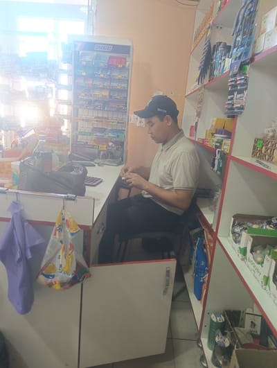 The image depicts an indoor scene, likely a small convenience store or kiosk, located in Bulungur, Uzbekistan. A man wearing a tan polo shirt and a black baseball cap with "NEWYORK" embroidered on it is seated at a counter, appearing to be in the process of making a transaction or counting something. He is looking down at his hands. In front of him on the counter are a calculator, a black plastic bag, and some small items he is holding. The counter is also cluttered with various goods.

To the left of the man, shelves are visible, filled with packaged snacks, including "Snickers" bars. A purple garment hangs on a hook to the left of the counter. To the right of the man, more shelves are stacked with a variety of products, including personal care items like soap bars and batteries. A cardboard box containing small, round items, possibly candy or individual servings, is also present on the right side shelves. The overall environment suggests a retail setting with a wide assortment of small consumer goods.