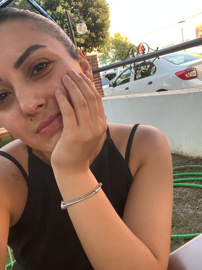 The image shows a close-up, selfie-style shot of a woman with her hand resting on her cheek. She has dark hair pulled back, dark eyes, and light skin with some freckles. She is wearing a black tank top and a silver bracelet on her right wrist. Behind her, a white car is visible parked on a street. The background also includes trees, a brick wall, and a decorative metal railing. The lighting suggests it is late afternoon or early evening. The image appears to have been taken outdoors, possibly in a residential or public area of Yunus Emre, Turkey, given the context.