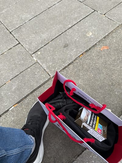 Viewed from a first-person, slightly downward perspective, a person's left leg and a shopping bag rest on a paved sidewalk in Leidschendam, Netherlands. The visible leg is clad in blue denim jeans, cuffed at the ankle, revealing a white sock and a black sneaker with a thick white sole. A faint red "HUGO" logo is discernible on the sneaker's tongue.
The shopping bag, branded "HUGO.COM" on its interior rim, is white externally with a distinct red inner lining and features red rope handles, one of which shows visible fraying. Inside the bag, folded dark fabric, consistent with clothing like trousers or a sweater, is visible. On top of this clothing item rests a pack of cigarettes.
The cigarette pack displays Dutch health warnings: "ROKEN VERSTOPT UW SLAGADEREN" (Smoking clogs your arteries), accompanied by a graphic image of a diseased foot/leg, and "Tabaksrook bevat meer dan 70 stoffen die kanker veroorzaken" (Tobacco smoke contains more than 70 substances that cause cancer). The brand name "Camel" is faintly visible.
The ground surface is a grey paved sidewalk constructed from rectangular concrete pavers, exhibiting a granular texture. Scattered on the pavement are several discarded cigarette butts with white filters, a small, dried reddish-brown leaf, and a single light-colored circular mark, possibly dried chewing gum. The lighting is even and diffused, suggesting an overcast sky.