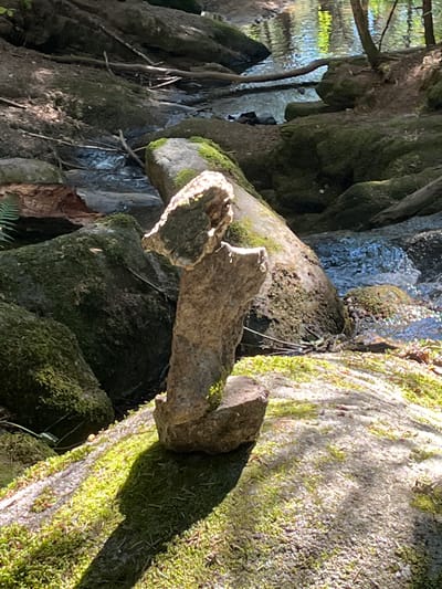 The image depicts a nature scene with a focus on a stacked stone formation in the foreground. The stones are weathered and covered in moss, with the dominant one appearing to be balanced vertically. The background shows a shallow stream flowing over rocks, with dappled sunlight filtering through the trees. The surrounding area is rocky and overgrown with moss and vegetation, suggesting a woodland or forest environment. The scene is set in a natural outdoor location, and it is not possible to determine the specific location within Gumières, France, from the visual information provided.