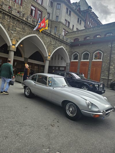 A silver vintage Jaguar E-Type is parked on an asphalt street in front of a stone building with arched doorways. The building has several windows with rounded tops and wooden doors. Three flags, one yellow with a blue emblem, one red with a white cross, and one blue and white, are hanging from the building. A dark-colored luxury car, possibly a Rolls-Royce, is parked behind the Jaguar. Two people are visible near the Jaguar. One person, wearing a brown sweater and dark pants, is standing and looking towards the car. Another person, wearing a green sweater and light blue jeans, is walking away from the camera, carrying two black bags. The sky is overcast. The scene appears to be in St. Moritz, Switzerland, as indicated by the flags.