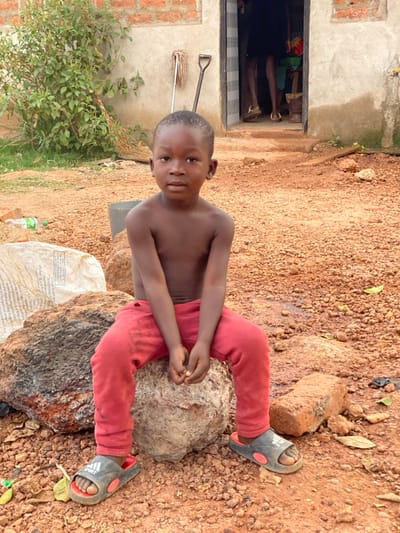A shirtless young boy wearing red pants and dark sandals sits cross-legged on a rock. He's positioned in a yard of reddish soil next to a small building with a doorway where other people are visible. A shovel and mop lean against the building wall. A bush is next to the building, and refuse is on the ground.