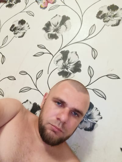 A man with a shaved head and a beard is lying down, looking at the camera. He is shirtless. The background is a wall with a floral pattern. There are flowers and leaves in black and white, and some colorful butterflies. The scene appears to be indoors, likely in a residential setting. The image does not provide any specific information about the location in Pishchane, Ukraine.