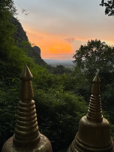 This is a landscape photograph taken at sunset from a vantage point overlooking a valley in Ban Khao Tang, Thailand. The foreground is dominated by two golden, stupa-like structures, one on the left and one on the right, which partially frame the view. The left stupa is closer to the camera and shows intricate ridged detailing on its upper section, tapering to a point. The right stupa is slightly further back and also exhibits a similar pointed top and ridged pattern.
Beyond the stupas, a dense expanse of lush green foliage, possibly bamboo and other tropical plants, fills the middle ground. The foliage appears to grow up a steep, rocky mountainside on the left, with a sheer rock face visible. A large tree with dark green leaves stands on the right side, also framing the distant landscape.
The background reveals a hazy, expansive valley with rolling hills and layers of vegetation receding into the distance. The sky above is a dramatic canvas of sunset colors, with a soft gradient from a pale blue-grey at the top to warm hues of orange and yellow near the horizon. A single, fluffy cloud is illuminated by the setting sun, adding a focal point to the sky. The overall impression is one of serene natural beauty and spiritual ambiance, likely a viewpoint from a temple or a place of worship. No people are visible in the image.