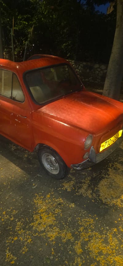 The image shows a close-up, rotated view of a small, red, vintage car parked on a paved surface. The car appears to be a Fiat 600 or a similar model from the mid-20th century. It has a distinctive rounded shape, with a single door visible on the driver's side. The front of the car features two round headlights and a black bumper. A yellow license plate with black text is attached to the front. The tire on the visible side is silver with a black sidewall. The car is parked in what appears to be an outdoor setting, with some greenery and a stone wall visible in the background. The ground is covered in scattered yellow petals, suggesting it might be spring or fall. The lighting indicates it is nighttime or dusk. There is no visible activity or people in the image. The overall impression is of a parked classic car in a dimly lit outdoor environment. The visible text on the license plate is "710-28R".