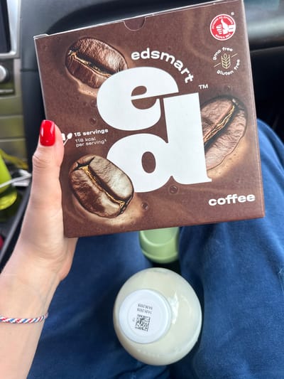 The image shows a person holding a box of "edsmart coffee 58". The box is brown with coffee bean images and labels indicating it's GMO-free and gluten-free. The box indicates that there are "15 servings 118 kcal per serving¹". Next to the box is a bottle with a white cap; text on the cap reads "14.05.2026 04.06.2026" along with a number "13". The person has red nail polish and is wearing a blue and red rope bracelet. The person's lap is covered with blue clothing. The background is blurry, but suggests the inside of a car with green objects. There is a Russian text that reads "Лучшие из Лучших" on the box, which is translated to "Best of the Best". There is no indication of this scene taking place in the city of Gornyi, Russia.