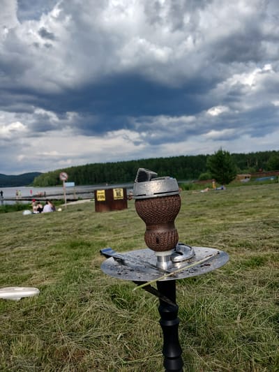 An outdoor scene captures a shisha pipe in the foreground, positioned on a grassy area. In the background, a body of water, likely a lake or reservoir, is visible, bordered by a forest. The sky is filled with dramatic, dark clouds, suggesting an impending storm or overcast weather. Several people are scattered across the landscape, some near the water's edge and others on the grass. A warning sign with a red circle and diagonal line is placed on a post to the left. Two metal containers, possibly bins, with Cyrillic lettering are situated on the grass behind the shisha pipe. The overall impression is a recreational setting on a beach or park, with a focus on the shisha pipe as the primary subject. The location is identified as the city of Revda, Russia.