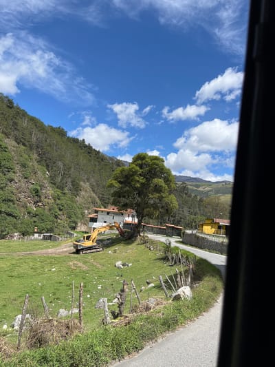 The image depicts a rural landscape in Mucurubá, Venezuela, under a bright blue sky with scattered white clouds. The scene features a steep, tree-covered mountainside on the left, leading down to a grassy field. In the field, a yellow excavator is parked near a white building with a red-tiled roof, labeled "POSADA." To the right of the field, a road curves past a yellow building with a red roof, bordered by a stone wall and white railings. A weathered wooden fence with sparse vegetation runs along the edge of the road in the foreground. The overall impression is of a serene, natural setting with signs of construction or development.